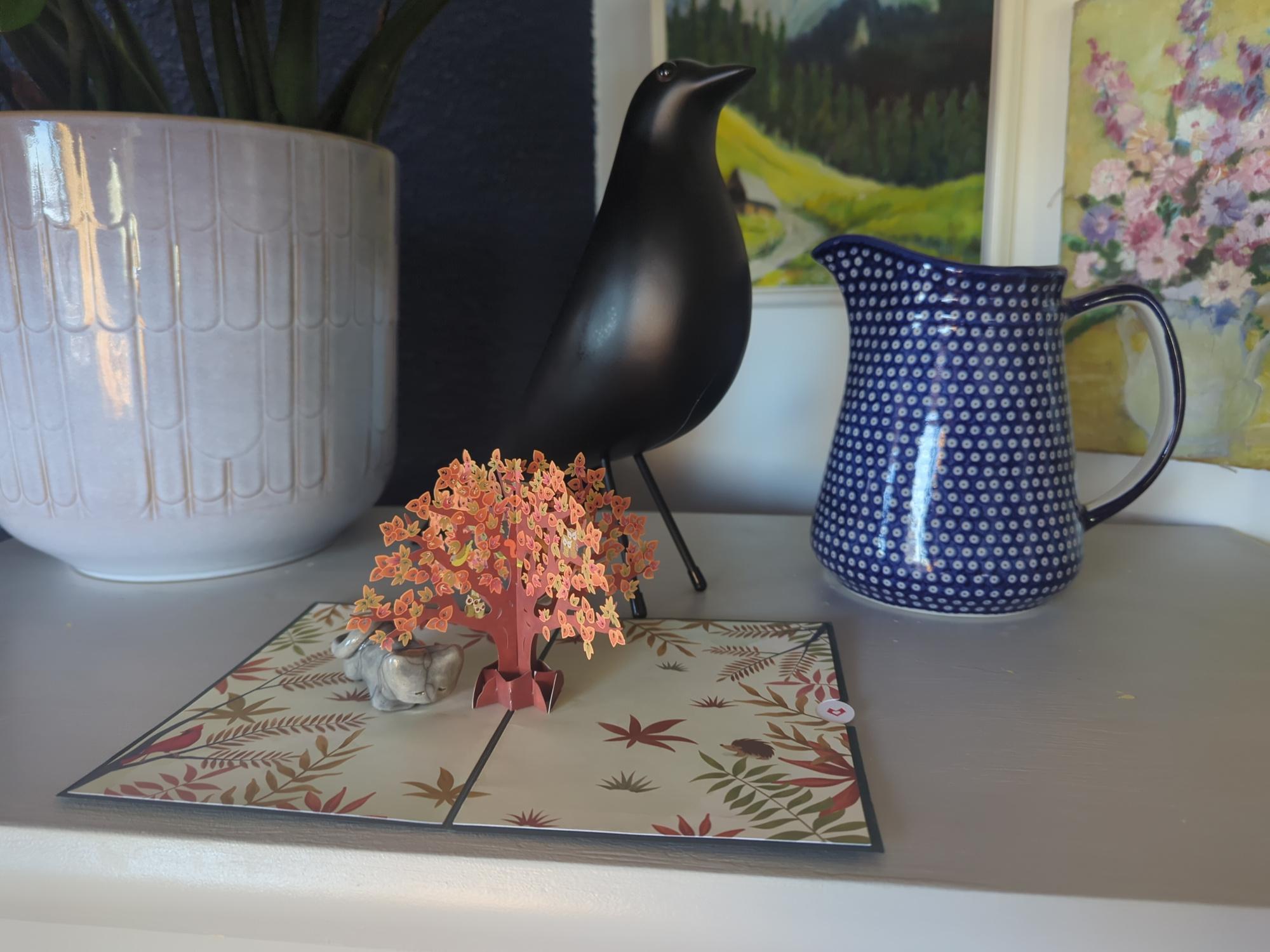 an open pop-up greeting card on a table, weighted down with a tiny ceramic cat. behind it is a vase, a potted plant, and a bird statuette.