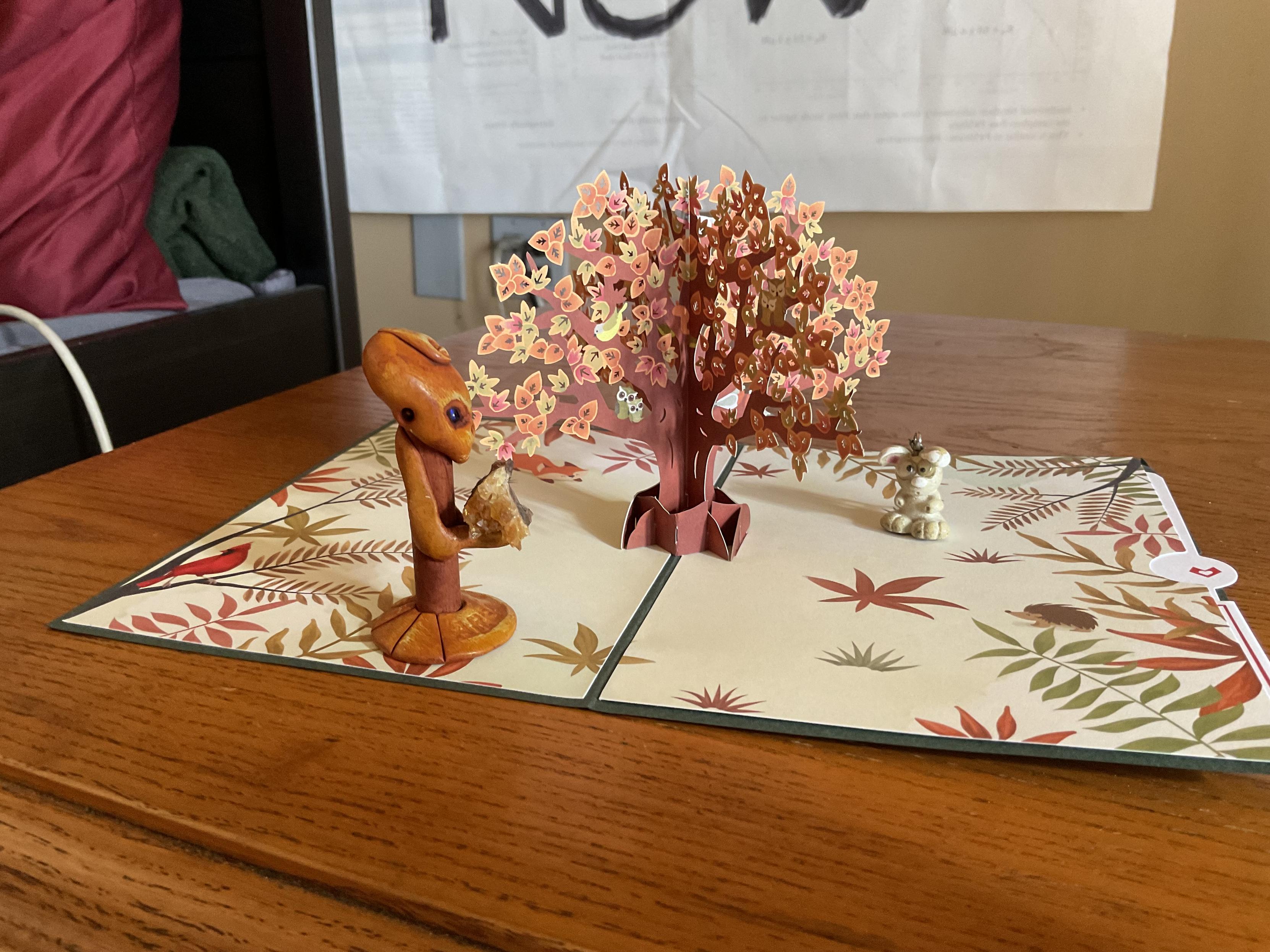 an open pop-up greeting card. the pop-up is an autumnal tree, and the inside of the card has autumn leaf designs. two small figurines sit on the card to hold it open flat.